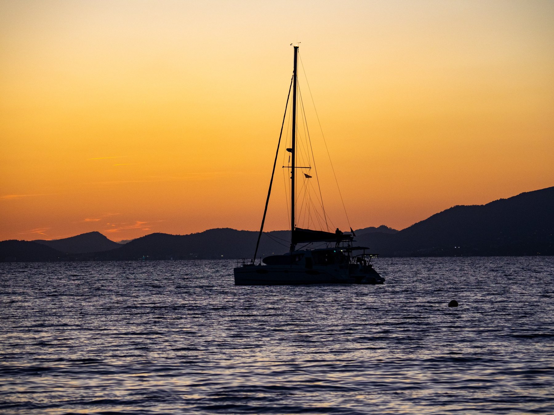 Silhouette eines Katamarans vor orangefarbenem Abendhimmel und dunklen Bergen am Meer